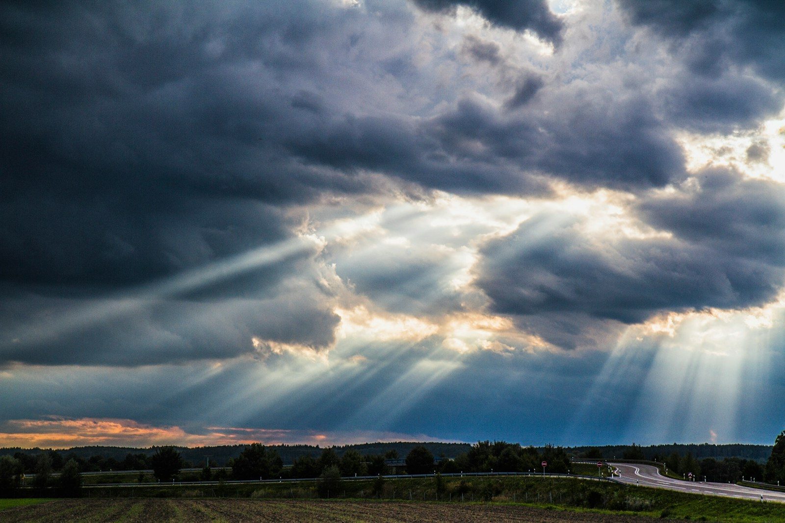 Sunlight streaming through dark storm clouds over a rural road and field, symbolizing the Holy Spirit
